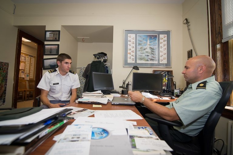 A cadet in a white uniform sits across from an officer in a green uniform in a cluttered office, engaged in conversation. The officer is seated behind a desk with papers, books, and computer monitors, while the cadet listens attentively.