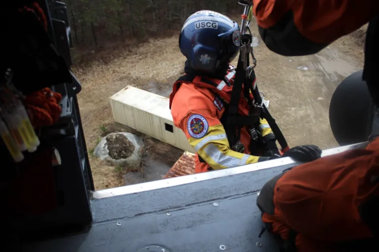 Coast Guard rescue crew member in blue helmet and orange survival suit being lowered from helicopter viewed from aircraft door.