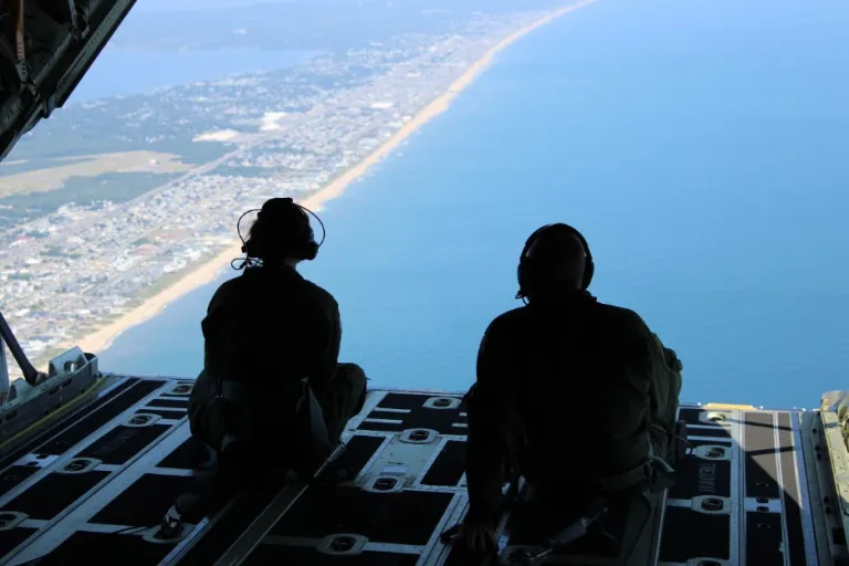 Silhouetted military personnel with headsets sitting at open aircraft ramp, overlooking coastline with beach and ocean visible from high altitude.