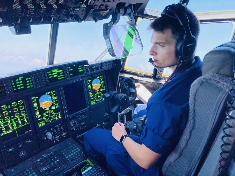 Pilot in blue flight suit operating aircraft controls in modern cockpit with digital displays and avionics screens showing flight data, clear blue sky visible through windows.