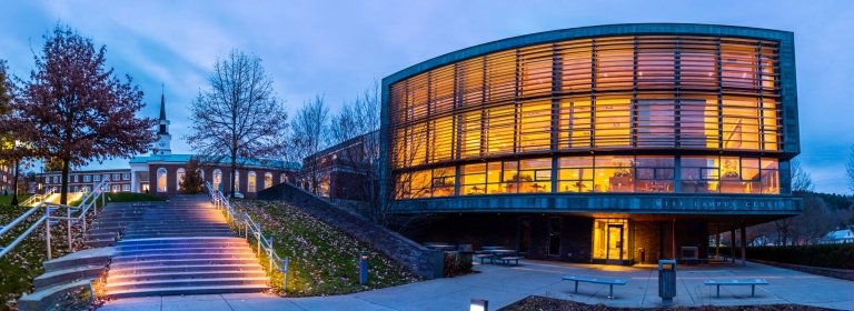 Evening view of Norwich University’s Wise Campus Center with its curved, glass-fronted facade glowing warmly, beside the illuminated steps leading to Plumley Armory and White Chapel.