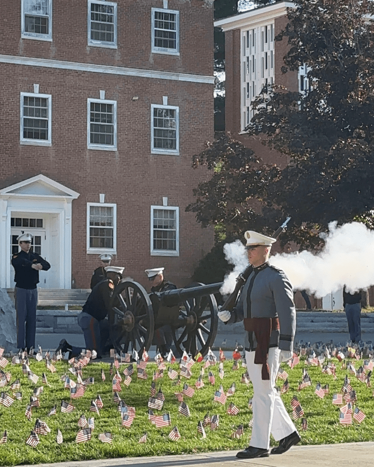 Ceremonial cannon firing by uniformed individuals in front of a brick building, surrounded by a field of small flags.