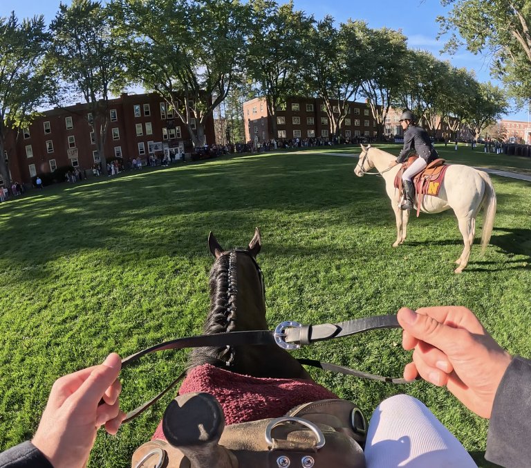 First-person view from horseback on a green parade field, with another rider on a white horse facing a crowd by brick dorms.