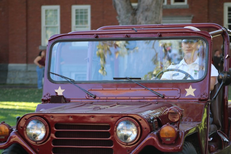 Cadet in uniform drives a vintage maroon military jeep past a brick campus building.