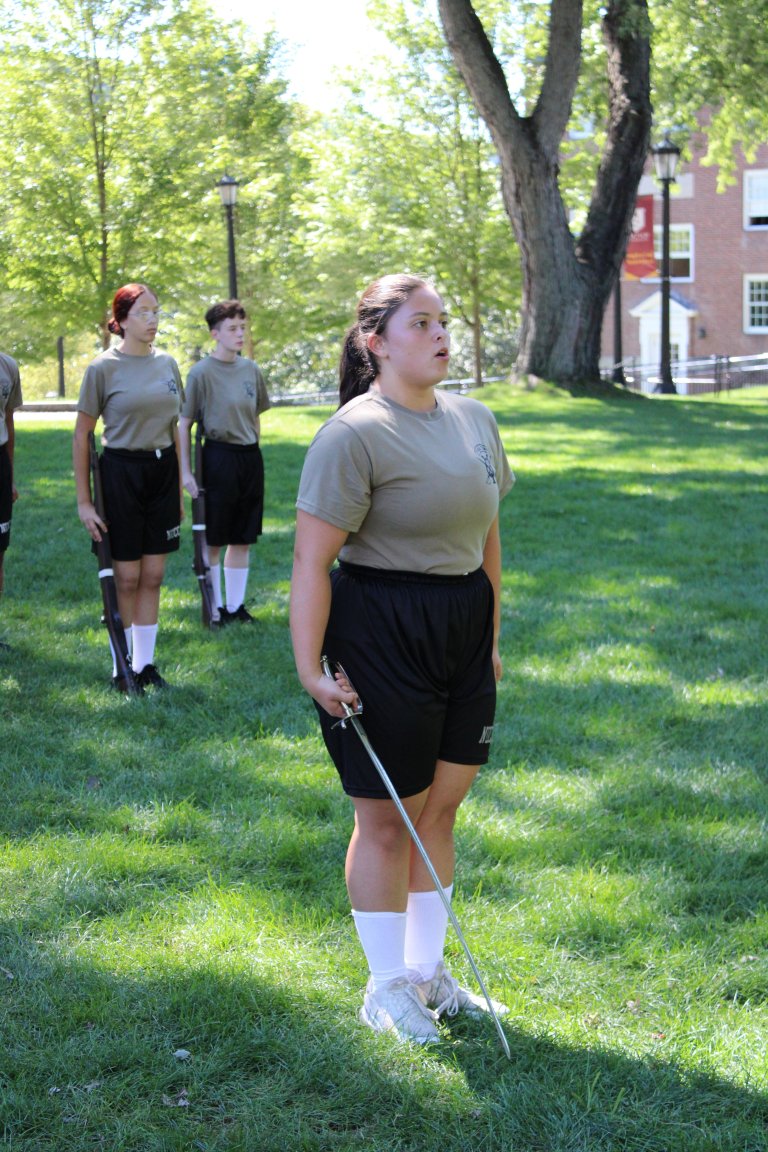 Students in uniform practice drill movements with swords on a grassy campus lawn.