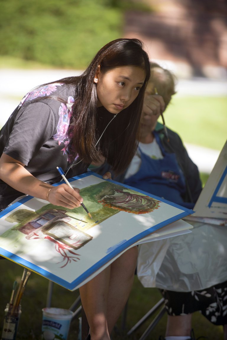 Student paints on a canvas outdoors while seated beside another person.