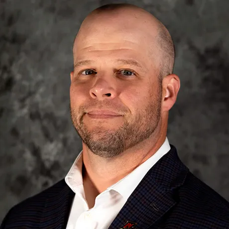 A man with a shaved head, trimmed beard, and wearing a dark plaid suit jacket over a white shirt, standing against a textured gray background.