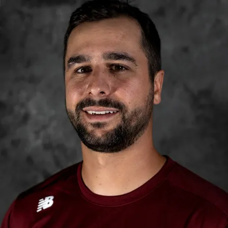 A man with short dark brown hair and a beard, wearing a maroon athletic shirt with a white logo, standing against a textured dark gray background.