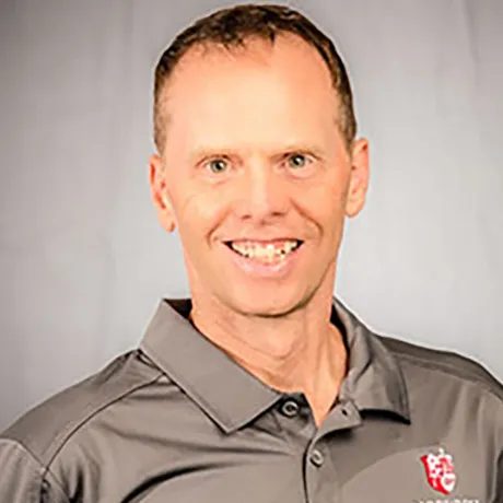 A smiling man with short brown hair wearing a gray polo shirt with a red and white logo, standing against a light gray background.