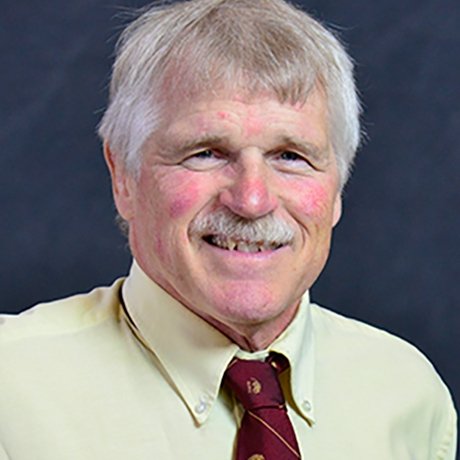An older man with light gray hair and a mustache, wearing a pale yellow dress shirt and a maroon tie with a subtle pattern, smiling in front of a dark background.