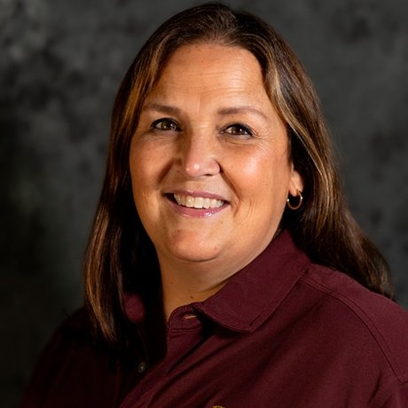 Smiling woman with straight, shoulder-length brown hair wearing a maroon collared shirt, posed against a dark mottled background.