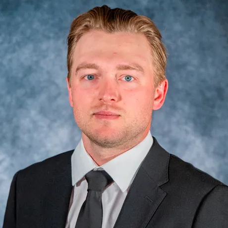 Professional head-and-shoulders portrait of a man wearing a dark suit, white shirt, and dark tie against a neutral blue-gray background.