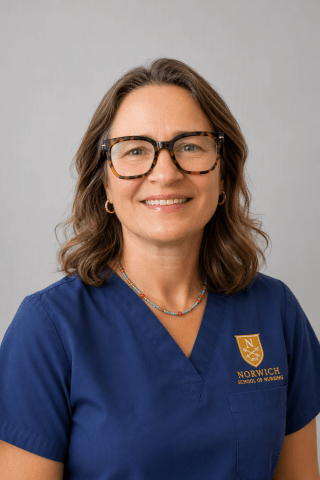 A person wearing glasses and blue Norwich University scrubs poses against a gray background.