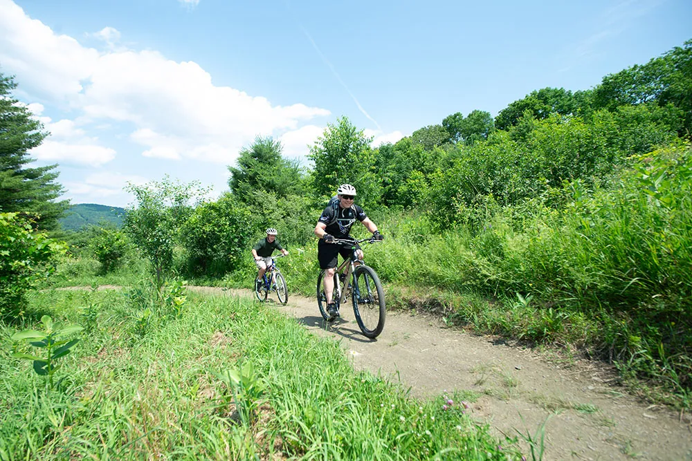 Two people ride mountain bikes on a dirt path through green, leafy trees under a blue sky with scattered clouds.