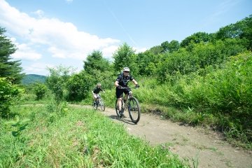 Two people ride mountain bikes on a dirt path through green, leafy trees under a blue sky with scattered clouds.