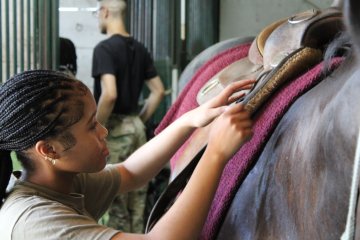 A person tightens the saddle on a horse while another stands in the background in a stable.