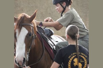 Cadet wearing a riding helmet adjusts the saddle while mounted on a horse, with an NU Cavalry Troop instructor standing beside them.