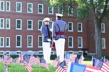 Two cadets in gray dress uniforms and white hats stand facing each other on a lawn filled with small American flags, with a red brick campus building in the background.