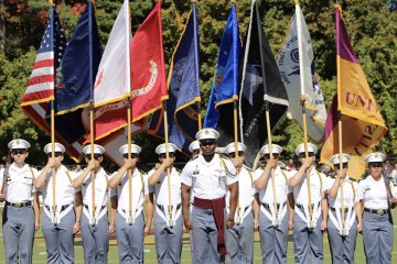 Full color guard line of cadets holds multiple U.S. and unit flags upright during an outdoor ceremony.