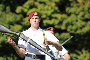 Cadet in a white uniform and red beret tosses and spins a rifle during a precision drill performance.