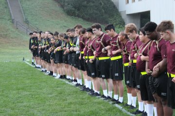 Long line of rooks in maroon shirts and black shorts stand at attention on a field, each holding a rifle.