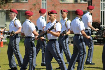 Squad of cadets in white shirts, gray pants, and red berets marches in step on a parade field while carrying rifles.