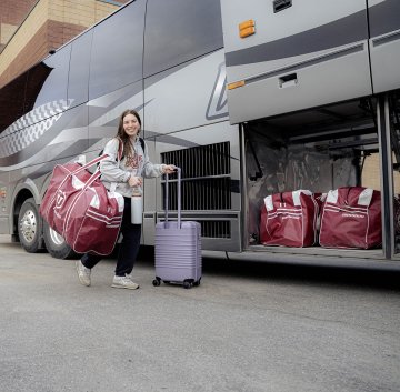 Norwich University women’s ice hockey player carries luggage and team gear beside bus while departing for NCAA Frozen Four 