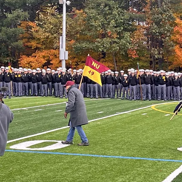 A person walks across a football field carrying a red and gold NU flag as uniformed cadets stand in formation behind them.
